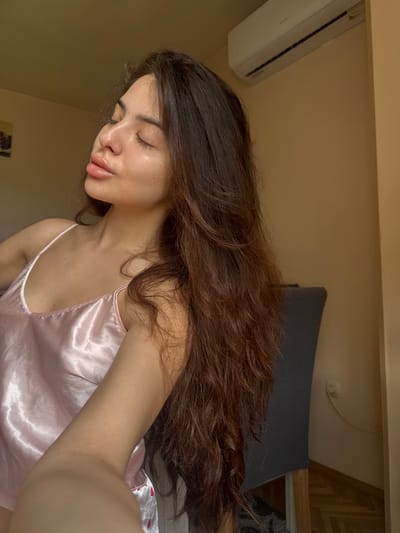 This indoor image, located in Plovdiv, Bulgaria, features a single young adult female captured from a chest-up perspective, indicative of a self-portrait. She has long, dark brown, wavy hair that flows over her shoulders and chest. Her eyes are closed, and her head is tilted upwards and to her left, suggesting a moment of repose with her face illuminated by an unseen light source. Her lips are full and slightly parted. She is wearing a light pink, satin-finish camisole with thin shoulder straps. Her right forearm is visible in the lower-left foreground, consistent with holding the camera.

The background consists of a plain, light brown wall. A white, wall-mounted split-unit air conditioner is affixed to the upper right section of this wall. A segment of a dark grey, high-backed chair is partially visible behind the subject on the right. On the right wall, a white electrical wall plate, possibly an outlet or switch, is present, from which a white insulated cable extends downwards and curves. The lower right portion of the scene reveals a polished wooden parquet floor. A framed object with indiscernible details is dimly visible on the upper left wall.