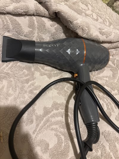 A gray hair dryer with a diamond pattern and a copper-colored ring around the barrel is lying on a beige towel with a raised floral pattern. The hair dryer's nozzle is pointed to the left. A black cord is attached to the hair dryer and is coiled around it. The scene is indoors, and no people are present. There is no information available about the location in Tbilisi, Georgia.