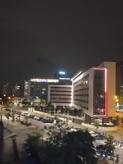 This image shows a nighttime view of an urban street scene in Madrid, Spain, likely in the vicinity of the IFEMA trade fair complex. The foreground features a street lined with parked cars, motorcycles, and a traffic sign. Several trees are visible, their dark foliage contrasting with the illuminated buildings.

In the mid-ground, two modern buildings dominate the scene. The building on the left is a multi-story structure with horizontal banding and numerous windows, some of which are lit. The building on the right is taller and features a striking facade with a large glass section, accented by a vertical red stripe and illuminated windows. The signage "Barceló Imagine" is visible on the taller building.

In the background, the cityscape continues with more buildings and streetlights. The sky is dark and cloudy, indicating nighttime. The lighting suggests a well-lit urban environment. No individuals are clearly discernible in the image, but the presence of parked vehicles indicates general activity in the area.