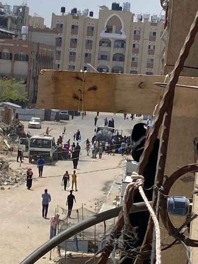 The image, oriented correctly, depicts an outdoor street scene in Khan Yunis, Palestinian Territories, captured from an elevated perspective. The central focus is a wide, unpaved or dirt street populated by numerous people and several vehicles. Long, distinct shadows of the individuals stretch across the ground, indicating a low sun angle, suggesting either early morning or late afternoon under a clear, bright sky.
People are seen standing in groups and walking along the street. Vehicles include a prominent white truck, possibly a utility or flatbed model, and at least two passenger cars. Along the edges of the street, especially on the right side, there are visible piles of rubble and debris, suggesting prior damage, ongoing demolition, or construction in the vicinity.
In the background, a prominent multi-story building with a light-colored facade stands out. It features multiple rows of windows, many of which have distinctive light-colored frames or shutters forming a 'K'-like pattern. A large arched entrance is discernible on a lower level of this building, and several dark water tanks are placed on its rooftop. An adjacent, darker building is also visible. The immediate foreground is obscured by blurred structural elements, wires, and possibly parts of a railing or another building, consistent with an elevated vantage point. No discernible text is visible on any objects or structures within the frame. The scene captures a moment of daily life with people congregating on a street amidst urban structures and scattered debris.