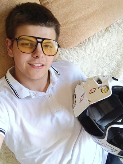 A young man with short, dark brown hair, wearing black-framed yellow-tinted glasses and a white polo shirt with black striped trim on the collar, is lying on a light-colored, furry surface, possibly a blanket or rug. He is holding a white and black helmet with stickers on it. A tan or beige colored pillow or cushion is visible in the upper left corner of the image. The location is inferred to be Andreyevo, Russia.