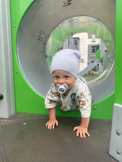 An infant is crawling on the ground inside a green playground tunnel. The infant wears a grey beanie and a light-colored shirt with a pattern of lions. A pacifier is in the infant's mouth. The tunnel is made of metal and plastic. Outside the tunnel, buildings are visible. The ground is a dark, textured surface with some white chalk marks.