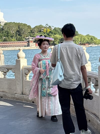 This image captures two individuals on a stone bridge or walkway beside a body of water in Xicheng District, China, likely during a photoshoot or tourist activity. The sky is bright and clear, indicating daytime with good weather.
In the foreground, a man is seen from behind, wearing a light-colored, short-sleeved collared shirt, black pants, and white sneakers. He has short dark hair and a light blue crossbody bag slung over his shoulder. He holds a black camera in his right hand, positioned as if taking a photo.
To his left and slightly in front, a woman stands, facing towards the camera. She is adorned in an elaborate traditional Chinese dress, possibly a hanfu, featuring layers of pink, light blue, and white fabric with intricate floral patterns. Her hair is styled traditionally, and she wears a decorative headpiece with purple flowers. She holds her hands gracefully, posing for a picture.
The setting includes a sturdy stone balustrade on the bridge, separating the walkway from a calm body of water, which reflects the light. Lush green trees and foliage line the opposite bank and extend into the background, with glimpses of traditional-style buildings beyond. The overall scene suggests a scenic historical park or garden. There is no visible text in the image.