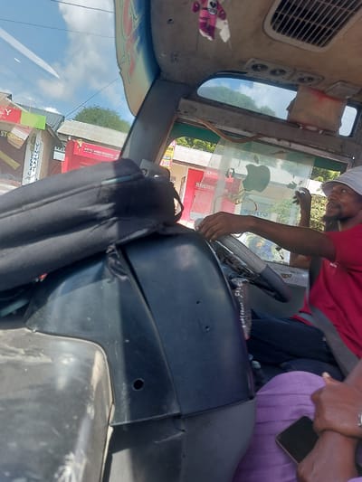 The image captures the interior of a vehicle, possibly a bus or van, with a man in a red shirt driving. He is wearing a white hat and has his hand on the steering wheel. A passenger in a purple shirt is seated next to him, holding a phone.
Through the windshield, the street scene is visible, showing commercial buildings with signs in Swahili. "Airtel Money Branch" is visible in red, and "STOORE" and "VINYWAJI" on storefronts. The general location appears to be Dar es-Salaam, Tanzania based on the language and style of signage.