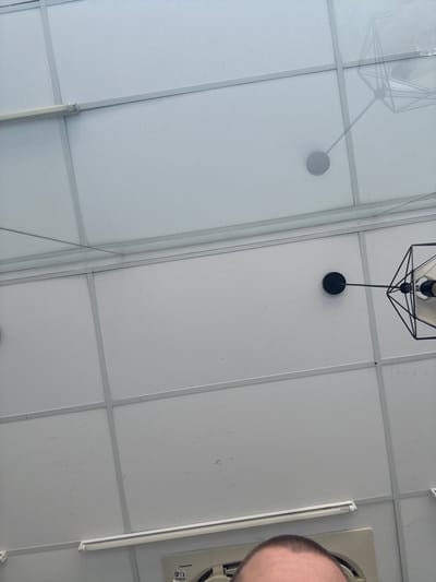 The image shows a ceiling with a grid of white panels, possibly a drop ceiling, with metal framing. There are small water droplets on the ceiling panels. Two light fixtures are visible. One is a long fluorescent tube light mounted horizontally along one of the metal beams. The other is a black geometric pendant light with a white shade, suspended from a black rod attached to a circular black base. A faint shadow of a similar geometric fixture is cast on the ceiling to the left. The image is taken from a low angle, looking upwards. The bottom of the frame shows the top of a person's head, a bald man with short brown hair. Below the man's head, part of a white electrical appliance with a Panasonic logo and some text is visible. The overall impression is that of an indoor space with a somewhat utilitarian ceiling. The provided context of Kuta, Indonesia, suggests this could be a commercial building, office, or public space in that area.