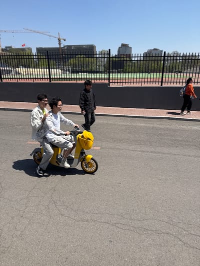 The image shows a scene in Zhongtun, China, on a sunny day. Two young men are precariously riding a shared yellow three-wheeled scooter on an asphalt road. The rider at the front wears a white shirt, a white jacket, and light-colored pants, while the passenger is dressed similarly. A third man in a black jacket stands to the side, holding a phone, possibly filming or photographing the scene. In the background, a fence separates the road from a sports field and some buildings, including what looks like a construction site with cranes. Another person in an orange top and black pants is walking in the background near the fence. The sky is clear and blue.