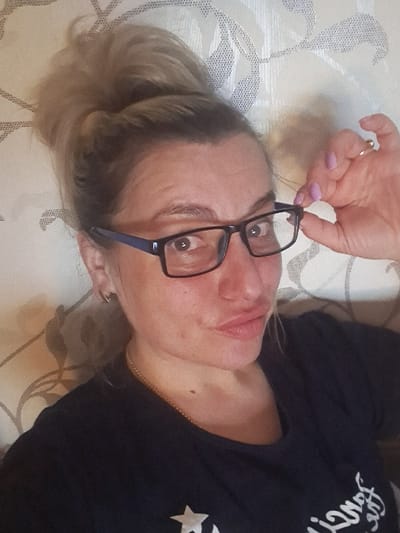 The specific location within the city of Yegoryevsk, Russia, cannot be determined from the visual information provided. The scene is set indoors against a light-colored background with a patterned, possibly floral or botanical, motif in a darker shade.

One adult female is depicted in a close-up, self-portrait orientation. She has blonde or light brown hair styled in an elevated bun on the top of her head. Her complexion appears fair with some visible redness or minor skin irregularities. Her eyes are wide open, directed towards the camera. Her lips are slightly puckered. She is wearing dark-framed eyeglasses with blue temples; her right hand is raised, with manicured fingers (light purple nail polish) gripping the right temple of the eyeglasses, suggesting an adjustment or removal action. A gold-colored ring is visible on the ring finger of her right hand. She is wearing a dark, likely navy or black, short-sleeved t-shirt, partially visible, which features a white star graphic on the lower left chest area. A thin gold-colored chain is visible around her neck, and a small gold-colored hoop earring is present in her left ear.