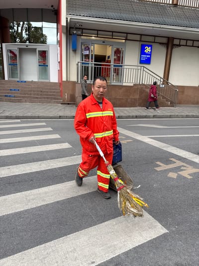 The image depicts a street scene in Jiangjin, China, featuring four individuals and two commercial buildings under ambient daylight.

The primary subject, a male of East Asian descent, is positioned slightly right of the frame's center, looking directly at the camera with a neutral expression. He wears a bright orange full-body uniform with yellow reflective stripes across the chest, arms, and lower legs, paired with dark shoes. In his right hand, he holds a broom with a white handle and natural bristles, secured with yellow and red tape. A dark grey dustpan is partially visible beneath the broom. A dark blue bag is carried over his left shoulder, resting against his left hip. He is captured in mid-stride, walking from right to left across a pedestrian crosswalk marked with white stripes on an asphalt road. Yellow Chinese characters are painted on the road surface to the right of the crosswalk.

In the background, two buildings are visible. The building on the right exhibits a traditional dark tiled roof, a brown lower facade, and light-colored upper walls with decorative wooden supports. Its entrance features automatic glass doors, accessed via a short flight of steps bordered by a metal handrail. Above the entrance, a blue rectangular sign displays "24" in white, accompanied by Chinese characters "自助银行" and the English text "Self-Service Banking." Red decorative elements are visible around the entrance. A second male, of East Asian descent, wearing a light brown jacket, stands partially obscured in the doorway of this building. Further to the right, a female, also of East Asian descent, wearing a dark red jacket and dark pants, walks on the sidewalk, partially obscured by the building's railing.

The building on the left features a modern design with a predominantly glass facade and a prominent red vertical structural column. It has a series of steps leading up to two sets of closed glass doors, behind which a white roller shutter is visible. Red festive decorations are present inside this building's entrance. Security cameras are mounted on the exterior of both buildings. The scene is evenly lit, indicating an overcast sky.