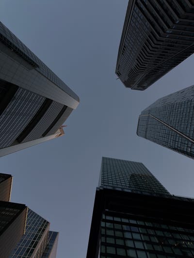 This media file is an upward-looking shot of several modern skyscrapers against a clear, pale blue sky, suggesting daytime. The composition creates a sense of looking up from ground level, emphasizing the immense height and architectural complexity of the buildings. The architecture varies, with some structures featuring dark, angular facades and others clad in glass with visible internal lighting, hinting at offices or apartments.
The scene appears to be a business district or a densely populated urban area, characteristic of a major city. The lighting and the absence of shadows suggest it might be late afternoon or early morning, though the overall lighting is even. There are no people or obvious vehicles visible, focusing the attention entirely on the architectural elements. The image conveys a feeling of scale and modernity. No visible text is present in the image. Based on the prompt's location context, this scene is likely in Frankfurt, Germany, a city known for its impressive skyline.