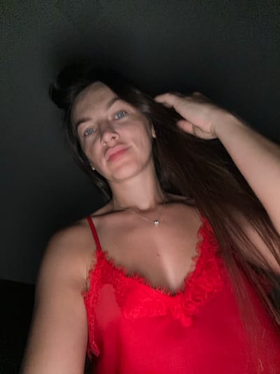 The image is a low-angle, close-up selfie of a woman with long, dark hair. She is wearing a red lace-trimmed camisole and a delicate necklace with a single pendant. Her left hand is raised, touching her hair. The background is dark and out of focus. The image appears to have been taken indoors, possibly in a dimly lit room. There is no indication of the location being Donji Pobori, Montenegro.