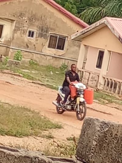 The image depicts a daytime scene in Adankolo, Nigeria, featuring an adult man riding a motorcycle on a reddish-brown dirt road. The man, with a dark complexion, short hair, and a beard, is dressed in a dark short-sleeved shirt and dark trousers, and is focused on driving.
His motorcycle is light-colored (white and yellow) and has two prominent, bright orange jerrycans attached – one at the front and another on the side, indicating the transport of liquids such as fuel or water. The road beneath him is unpaved and somewhat uneven, with patches of green grass and weeds growing along its edges.
In the background, two houses are visible. The structure on the left is light brown with areas of peeling paint and features several dark-framed windows, topped with a reddish roof. To its right, another house, lighter in color (possibly pinkish-beige) with a similar reddish roof and a visible dark-framed window, is partially seen. Both houses appear aged. Lush green foliage, including what looks like palm fronds, is present behind and between the buildings. In the lower right foreground, a large, textured grey concrete or stone block rests. The lighting is soft and diffused, suggesting either an overcast sky or ambient daylight. No visible text is discernible in the image.