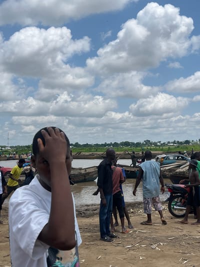 A group of people are gathered near a river in Nemnei, Nigeria. In the foreground, a person in a white shirt has their hand on their head. In the background, several boats are docked along the riverbank, and a few vehicles, including a car and a motorcycle, are visible. The sky is filled with scattered clouds.