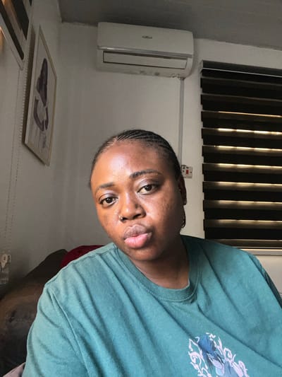A medium shot captures a young Black woman from the chest up, posed in a selfie-style frame. She has braided hair and wears a green t-shirt with a graphic print on the front. Her facial expression is neutral, with her lips slightly pursed. In the background, a white air conditioning unit is mounted on the wall above horizontal blinds. A framed picture of a person is visible on the left wall. The scene appears to be indoors, likely in a residential setting, possibly within a city environment like Abuja, Nigeria, given the presence of typical urban interior decor.