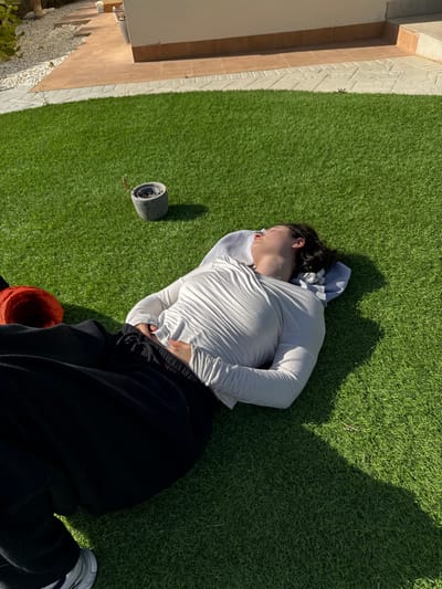 This image depicts an outdoor scene located in Fuente Álamo de Murcia, Spain. A single individual is positioned horizontally on a vibrant green artificial grass lawn, lying on their back with eyes closed, indicative of rest or sleep. Their head rests on a folded light-grey fabric item, possibly a towel. The person is dressed in a long-sleeved light-colored (possibly white or light grey) shirt with visible horizontal texture and black drawstring pants. Their left hand rests on their lower abdomen, while the right arm is extended downwards and partially obscured. A small portion of a light-colored athletic shoe is visible at the bottom left. To the left of the individual's head, on the artificial grass, is a small, grey, stone-like cylindrical planter containing a single sparse, dry plant stalk. Partially visible near the individual's left hip is an orange object, possibly a piece of clothing or fabric. Behind the artificial grass, a tiled patio or walkway with light-colored grout lines extends, bordered by a building wall featuring a reddish-brown base and a cream or light-beige upper section. The scene is illuminated by strong, direct sunlight, casting distinct shadows on the grass, notably to the right of the person and from the small planter.