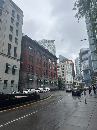 This media file captures a street scene in Vancouver, Canada, on an overcast day, possibly in the morning or afternoon, given the gray sky. The image is shot at an angle, looking up towards a cluster of buildings and down a wet street.

The scene features a mix of architectural styles. In the foreground and mid-ground, there are older brick buildings with arched windows and stone accents. Adjacent to these are more modern buildings with glass facades and angular designs, reaching high into the sky.

Several vehicles are visible on the wet asphalt road, including a black SUV and a line of white vans. The road also shows tire marks, suggesting recent activity. A yellow line demarcates a lane on the road. A few pedestrians can be seen walking on the sidewalk in the distance. A street lamp is also present in the composition.

The wet street and overcast sky indicate that it is raining or has recently rained. There are no strong indicators of a specific time of day other than the general subdued lighting. The emotions conveyed are neutral, focusing on the urban environment and its inhabitants. No significant text is legible in the image, though some small signs might be present on the vehicles or buildings, they are too indistinct to be read.