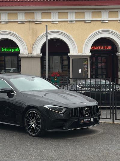 A black Mercedes-Benz sedan is parked on a wet asphalt street in front of a building with arched entrances. Above the entrances, neon signs read "IRISH PUB" in green and "ARAT'S PUB" in red. The building appears to be a commercial establishment, possibly a pub or bar, with dark windows. A black metal fence separates the street from the building's entrance area. A person is visible through the glass door on the right side of the "ARAT'S PUB" entrance, wearing a red garment. The sky is overcast, suggesting a cloudy or rainy day. The image appears to be taken in Ufa, Russia, as indicated by the license plate on the car which is typical for that region.