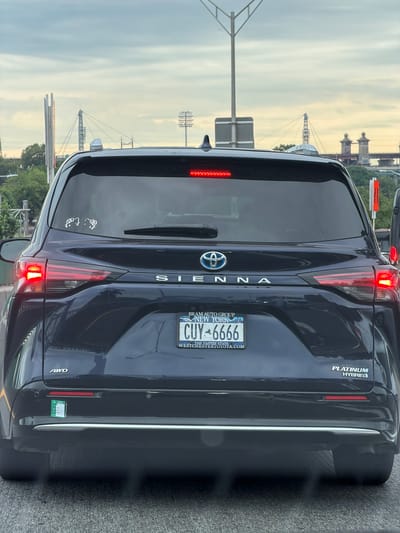 The image shows the rear of a dark blue Toyota Sienna minivan. The minivan has a New York license plate that reads "CUY-6666". The vehicle is on a road in what appears to be a city environment, possibly New York, given the license plate. In the background, there are bridges and other urban infrastructure, suggesting a location within or near New York City, United States of America. The sky is overcast with clouds. There are no people visible in the image. The minivan has an "AWD" badge on the lower left side of the bumper and a "PLATINUM HYBRID" badge on the lower right side. There are two small white stickers on the rear window, depicting cartoon figures.