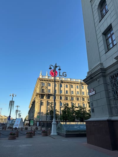 This is a street-level photograph taken in Minsk, Belarus, on a clear, sunny day. The image is oriented vertically, with the sky dominating the left side and the street and buildings on the right. The scene depicts an urban environment with a focus on architecture and street elements.
Prominently featured is a tall, ornate building with a classical facade, characterized by rows of windows, decorative detailing, and a projecting cornice. Adjacent to this building, on the left, is a portion of another, lighter-colored building with a more modern, gridded architectural style.
Several streetlights are visible. One notable lamppost has a circular red sign with a white symbol, possibly indicating a restriction or informational point. Another lamppost is larger and more industrial-looking, with several lights. In the foreground, planters with red flowers line the sidewalk, separated by chains.
Street signs and advertisements are present. One sign on the building's facade reads "20" and some text in Cyrillic, which appears to be a street name. A large digital billboard is visible further down the street, displaying colorful graphics and text, also in Cyrillic. The street itself is paved with gray bricks, and cars are visible in the distance, suggesting ongoing traffic.
The overall impression is one of a well-maintained urban street scene on a bright day. The clear blue sky and the sharp shadows cast by the buildings indicate it is likely midday. The architecture suggests a location in a European city, confirmed by the provided location context of Minsk, Belarus. There are no people clearly visible in the frame. The atmosphere appears calm and orderly.
