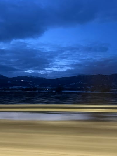 A landscape displays a contrast between cool and warm tones. The left side features a dark blue, cloudy sky meeting a dark, mountainous terrain beside a body of water. Light reflects subtly on the water's surface. The right side of the image is dominated by vertical lines of yellow or beige, creating a stark contrast with the natural elements on the left.