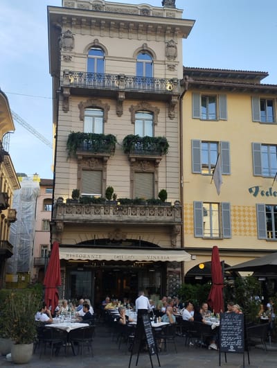 The image captures a street scene in Lugano, Switzerland, featuring the facade of a building housing "Ristorante Sass Cafe Vineria." The building is of classical architecture with ornate detailing on its upper floors, including balconies with wrought-iron railings and decorative stonework. The facade is a light beige color. Adjacent to it is a building painted a vibrant yellow, with grey shutters on its windows and a diamond-patterned design below the upper windows. A flag hangs limply from one of the yellow building's windows.
On the ground level, an outdoor seating area for the restaurant is set up. Several tables covered with white tablecloths are arranged on the cobblestone pavement. Diners, dressed in casual summer attire, are seated at these tables, engaged in conversation and enjoying meals. A waiter in a white shirt stands near a table on the left side of the frame. Two large, vibrant red umbrellas provide shade over some of the seating areas. Green bushes and potted plants add a touch of nature to the scene. In the background, a construction crane is visible against a clear blue sky, suggesting ongoing development in the city. The overall atmosphere is one of relaxed outdoor dining on a pleasant day.