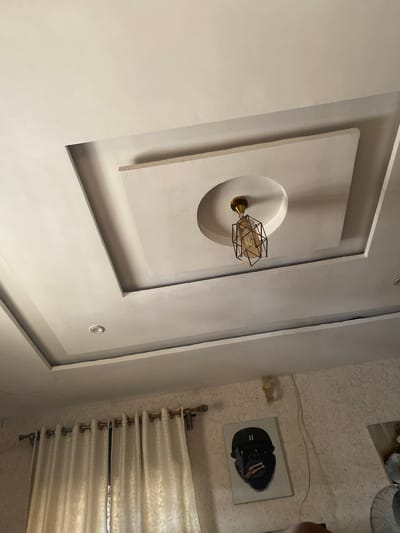 This image captures an indoor scene, likely a living room, with a focus on the ceiling and a portion of the wall. The ceiling is decorated with a modern, recessed design featuring a central light fixture. The fixture itself is geometric, with a gold-colored frame and a clear glass bulb visible inside. Adjacent to the ceiling feature, a textured wall is visible, adorned with a framed artwork. The artwork depicts a stylized portrait of what appears to be a chimpanzee or a gorilla, rendered in a dark, somewhat abstract style. Below the artwork, a section of off-white curtains is visible, drawn across a window. The lighting suggests it is daytime, with natural light filtering through the curtains. The overall impression is one of contemporary interior design with a touch of artistic flair. There is no discernible text within the image. The image appears to be taken in Giginyu, Nigeria.