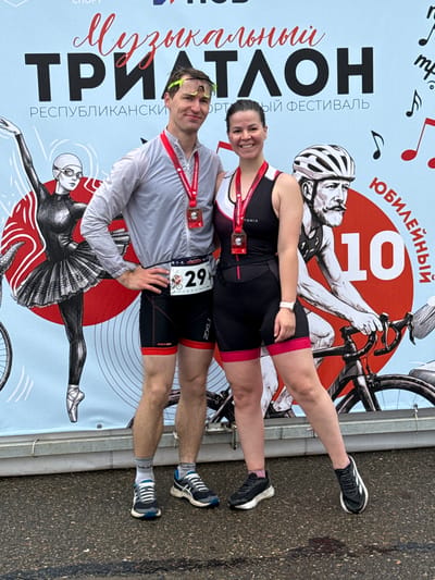 A man and a woman, appearing to be a couple, pose for a photo in front of a large, light blue banner. The banner features text that appears to be in Russian, with the word "ТРИАТЛОН" (Triathlon) prominently displayed in large black letters. Musical notes and illustrations of a ballerina and a cyclist are also visible on the banner.

The man, on the left, is wearing a light gray athletic jacket over a red and white medal lanyard. He has dark hair, a mustache, and is wearing yellow sports goggles on his forehead. He is wearing black athletic shorts with red trim, gray athletic socks, and dark blue and white athletic shoes. A race bib with the number "29" is attached to his shorts.

The woman, on the right, is wearing a black and pink athletic triathlon suit. She has dark hair pulled back and is also wearing a medal lanyard. She has a white smartwatch on her left wrist. She is wearing black athletic shoes with white accents and light pink socks.

They are standing on a wet, dark asphalt surface, possibly after a race, as indicated by their athletic attire and the medals. The overall scene suggests an outdoor event, likely a triathlon, in Votkinsk, Russia.