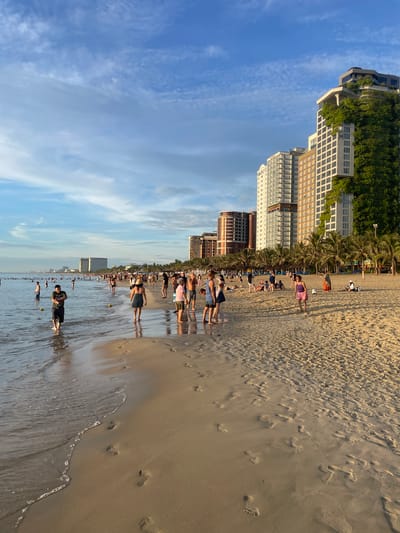 The image depicts My Khe Beach in Đà Nẵng, Vietnam, at golden hour. Numerous people are spread across the sandy beach and wading in the shallow ocean water. In the background, several tall, modern buildings line the coast, with one particularly striking building covered in lush green vegetation. Palm trees are visible between the buildings and the beach. The sky is a vibrant blue with scattered clouds, illuminated by the setting sun, casting warm light and long shadows across the scene. People are engaged in various activities such as walking, standing, and relaxing. Footprints are clearly visible in the wet sand near the water's edge.