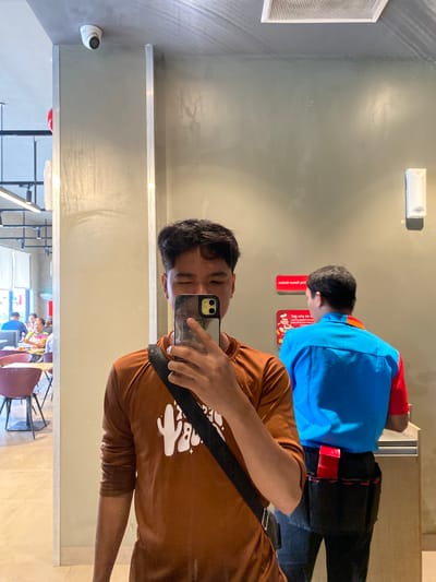 A young man, wearing a brown long-sleeved shirt with a white cactus graphic and a black crossbody bag, takes a selfie in a restaurant. He is standing near a wall with a mirror reflecting his image. In the background, visible through a large window and in the mirror's reflection, is a dining area with tables and chairs, and several people eating. Another person, wearing a blue and red uniform, is also visible in the reflection, standing near a counter. The environment appears to be a casual dining establishment, likely a fast-food restaurant, in Davao City, Philippines, indicated by the presence of a prominent red and white sign with a Jollibee mascot logo visible in the reflection. The lighting is bright, suggesting daytime. A security camera is mounted on the ceiling in the upper left portion of the image.