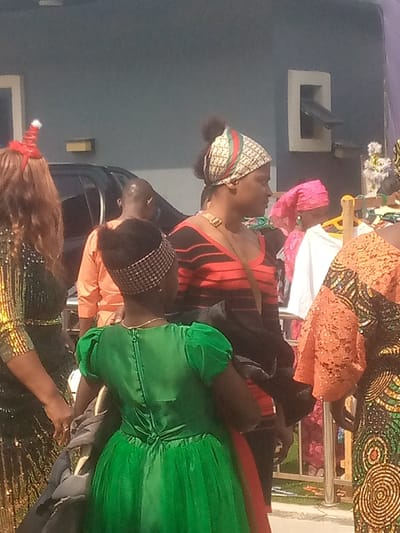 A group of people are standing outside. A young girl wears a green dress and checkered headband. A woman stands beside her, wearing a red and black striped top and a patterned head covering. Other people are visible, including a person in an orange shirt and another wearing a shiny green dress and a Santa hat headband. Clothes hang on a rack in the background. A building is visible behind the group.