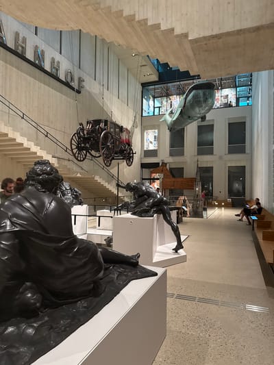 This image captures a wide-angle view of an indoor exhibition space, possibly a museum or gallery in Vienna, Austria. The scene is filled with a variety of sculptures and exhibits, creating a dynamic and visually rich environment.
In the foreground, a large, dark bronze sculpture of a reclining figure dominates the lower right of the frame. Its detailed musculature and dramatic pose suggest a classical or mythological theme. To the left and slightly behind this sculpture, another bronze figure is depicted in a more active, wrestling pose, reaching out towards something unseen.
Above these sculptures, a large model of a whale hangs suspended from the ceiling, its dark, metallic form casting shadows. Further back, a horse-drawn carriage with ornate detailing is also displayed, seemingly at a different height or angle. In the background, the architecture of the space is visible, featuring modern, clean lines with exposed concrete and wooden elements. Large windows reveal glimpses of other exhibits or parts of the museum.
A few people are visible in the scene, scattered throughout the exhibition. Two individuals are seated at computers on a raised platform in the distance, suggesting a digital interactive exhibit or a workspace. Other visitors can be seen walking through the space, interacting with the displays or observing the art.
The lighting appears to be a mix of artificial lights from the ceiling and ambient light filtering through the windows. There is no indication of the time of day or weather. The overall emotion evoked is one of awe and intellectual curiosity, characteristic of a museum visit.
Visible text includes individual letters spelling out words, such as "THE" and "ZEN," mounted on a vertical surface on the left side of the image. These might be part of a larger installation or exhibit title.