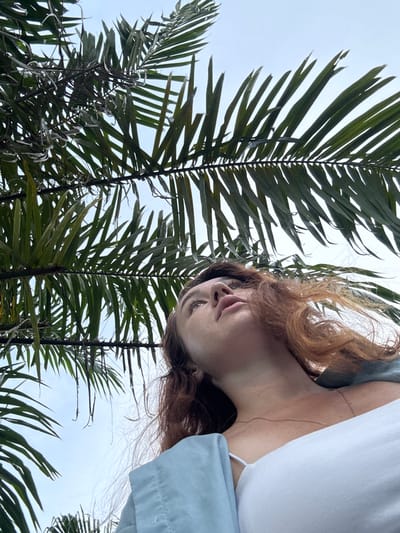 This is a low-angle, upward-looking shot of a young woman in Phuket City Municipality, Thailand. She has reddish-brown hair that is windblown and appears to be looking up and to her left. Her face is partially obscured by palm fronds, with only her nose and one eye visible. She is wearing a light blue garment with a white garment underneath, and a delicate necklace. The background is a cloudy sky, suggesting it is either early morning or late afternoon, or an overcast day. The palm fronds dominate the foreground, framing the woman's face and adding a tropical element to the scene. The overall mood is contemplative or perhaps a moment of observation, with the natural elements interacting with the human subject. There is no visible text in the image.