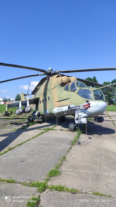 An olive drab and tan camouflage-painted attack helicopter is parked on a concrete pad under a clear blue sky. The helicopter has its main rotor blades extended, with the front rotor visible in the foreground. It is equipped with a chin-mounted Gatling gun and a series of rockets mounted on its wings. The helicopter is secured with rope barriers and stakes, suggesting it is on display at a museum or exhibition. The surrounding area consists of a paved surface with patches of green grass growing through cracks. In the background, a line of trees can be seen, and a white sign with red lettering is partially visible on the left. The image is dated 2026/7/15 10:21.
