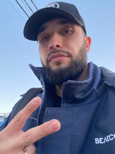 A selfie taken from a low angle features a man with a dark beard and a black baseball cap. He is wearing a navy blue jacket with white lettering reading "BEAECO" on the right side. The man is holding up two fingers in a peace sign. The background includes a blue sky and what appear to be power lines. Based on the location provided, this image was taken in Koskolovo, Russia.