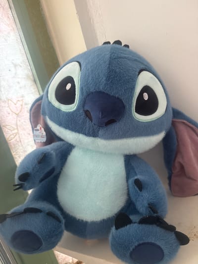 A stuffed toy resembling the character Stitch from Disney's Lilo & Stitch is sitting on a ledge. The toy is predominantly blue with a lighter blue stomach area, large black and white eyes, and dark blue accents on its nose, ears, and paws. It is positioned in front of a white wall and a window with a patterned curtain. A small, circular sticker is attached to the inside of the toy's right ear, featuring a drawing of a cloud and writing in Hebrew. The scene appears to be indoors, possibly in a residential setting in Herzliya, Israel, given the context provided.