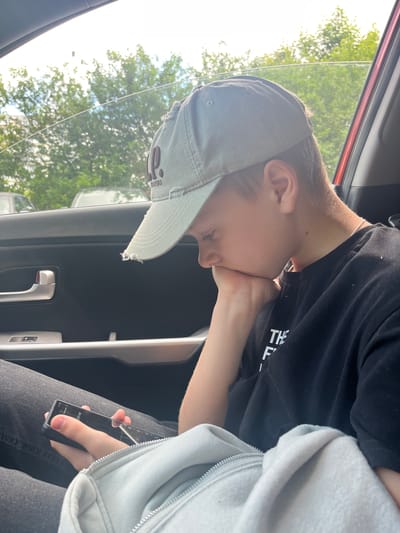 A young Caucasian male wearing a light-colored baseball cap and a black t-shirt is sitting in the passenger seat of a car. He is holding a smartphone in his right hand, with his chin resting on his left hand. The car's interior is visible, including a black door panel with a silver door handle and window controls. The background shows trees and other vehicles through the car window, suggesting an outdoor setting, possibly a parking lot. The overall lighting indicates it is daytime. The scene is captured from a medium close-up, focusing on the boy and his immediate surroundings within the car. There is no visual information in the image that can determine the location as Kaliningrad, Russia.