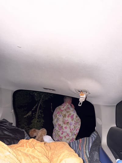 The image shows the interior of a vehicle at night, looking out an open rear window into darkness with some foliage visible. In the foreground, on the floor of the vehicle, are various items including orange towels or blankets, a black bag, and a sleeping bag with a colorful striped pattern. A person wearing a floral patterned shirt is sitting facing away from the camera, looking out the window. A small, light brown dog is standing between the person and the towels. Above the person, a white seatbelt buckle is attached to the ceiling of the vehicle. The scene appears to be set up for camping or sleeping inside the vehicle, likely in a natural or semi-natural environment. There is no indication of the specific location within the city of Vyshhorod, Ukraine.