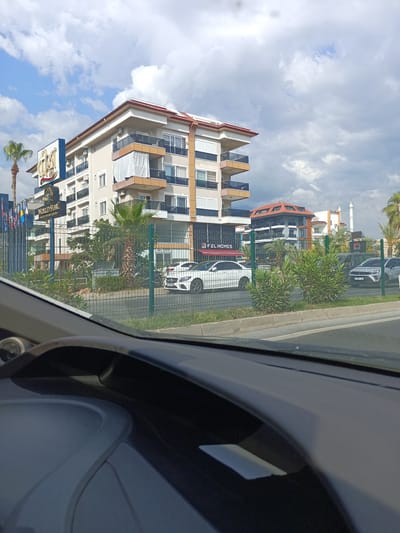 The image depicts a street view in Alanya, Turkey, on a cloudy day. In the foreground, the interior of a car is visible, showing the dashboard and windshield. Outside the car, a multi-story building, likely an apartment complex or hotel, dominates the scene. This building features a light-colored facade with balconies, some with white curtains. A prominent sign with "WORLD STAR - BEACH RESORT ESSE" is visible on the left side of the building, along with several flags. Adjacent to the building, another structure with an unusual angular roof design is present. In front of the buildings, a green metal fence separates the property from the street. Several cars are parked or driving on the street, including a white Mercedes-Benz. Lush green foliage and palm trees are interspersed around the buildings and along the street. A minaret of a mosque is visible in the background on the right. The overall environment suggests a sunny but partially overcast day in a developed urban area.