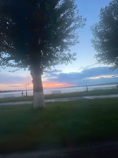 This image depicts a scenic view of the Votkinsk reservoir in Votkinsk, Russia, during sunset. The sky is painted with hues of blue, pink, and orange, transitioning into a clear blue overhead. Silhouetted trees frame the scene, their branches casting shadows on the foreground. A paved walkway runs along the reservoir, bordered by a decorative metal fence. A lamppost stands on the walkway, illuminated by the fading light. A lone individual is visible walking on the pathway. The foreground features a grassy area.