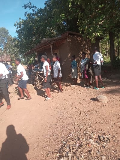 The image shows a group of eight people standing in front of a small, mud-brick structure. The structure is partially obscured by trees and has a thatched roof.  The individuals are all wearing casual clothing, with several wearing school uniforms. The people are gathered in front of the structure, and some appear to be interacting with each other. The ground is dusty and there is a pile of debris in the foreground. The image is likely taken in Malava, Kenya, at the coordinates provided (latitude: 0.53, longitude: 34.813). The image appears to have been taken on a sunny day, and the shadows cast by the individuals are visible on the ground.  The image is slightly blurred and has low resolution.