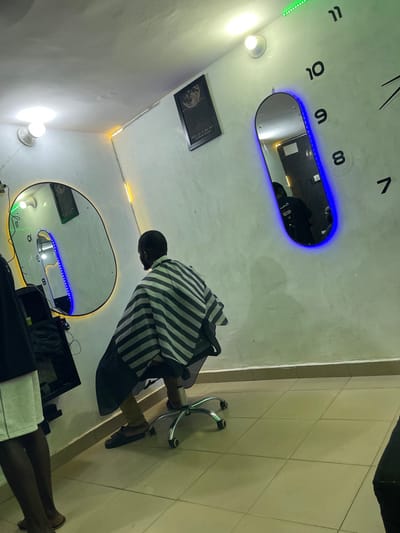 The image depicts the interior of a barbershop located in Unguwar Gabas, Nigeria. The scene shows a male individual seated in a barber's chair, viewed from behind, wearing a black and white striped barber's cape over brown trousers and dark slippers. His hair is short on the back of his head. The barber's chair has a chrome-plated base with five castor wheels.
The room features light-colored walls and a tiled floor. Multiple light sources illuminate the space, including two white ceiling lights, a green LED strip light along a ceiling edge, and yellow LED strip lighting along a wall corner. Two mirrors are visible: one large, irregularly shaped mirror on the left with a yellow LED frame, and one smaller, pill-shaped mirror on the right with a blue LED frame.
Reflected in the right pill-shaped mirror is another individual, likely wearing a dark top. On the wall, above the mirrors, is a framed picture of a full moon and large black numerical clock markings (7 through 11) directly applied to the wall. A dark-colored multi-tiered cabinet or shelf is partially visible to the left of the seated client. A third individual's lower legs and feet, wearing dark sandals and light shorts, are partially visible on the far left.