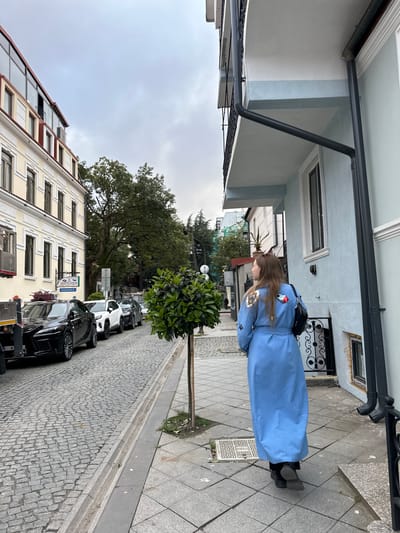 A woman wearing a long blue coat with embellishments on the back and sleeves is walking on a sidewalk in Batumi, Georgia. The sidewalk is tiled with grey square tiles. Next to the sidewalk is a cobblestone street lined with parked cars, including black and white vehicles. A neatly trimmed round tree stands between the sidewalk and the street. Buildings with multiple windows are visible on one side of the street, and a light blue building is on the other side. The sky is overcast. There is a sign with the word "INSURANCE" on one of the buildings.