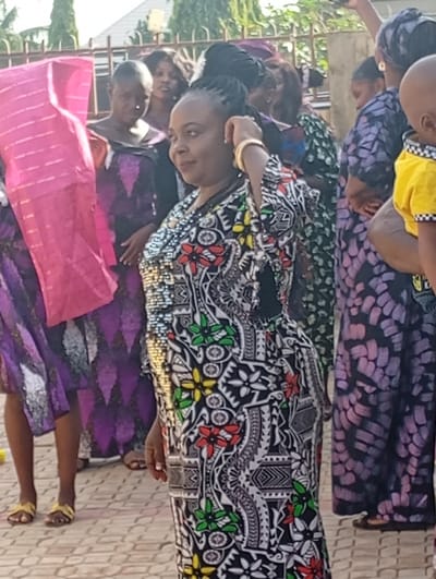 A medium-shot depicts a group of women standing outdoors on a paved surface, likely a public space in Rido, Nigeria, judging from the clothing styles and general environment. The central figure is a woman in a black and white floral dress with red, green, and yellow accents. She has dark hair styled in braids and is touching her hair with her right hand, while wearing a gold bracelet on her wrist. Behind her are several other women in patterned dresses, including purple, green, and pink fabrics. Some of the women are partially obscured. A child in a yellow shirt with a black and white patterned collar is visible to the right. In the background, there is greenery, possibly trees or bushes, and a partial view of a building. The lighting suggests it is daytime.