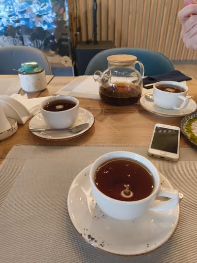 A high-angle, close-up shot depicts a table setting with three cups of tea, a glass teapot, a sugar dispenser, a cell phone, and a small plate of food. The table is covered with two placemats, one textured beige and the other a lighter, woven material. The background features a blurred interior with wooden paneled walls and upholstered chairs, suggesting a restaurant or cafe. A hand is visible in the upper right corner, making a gesture. The lighting is natural and diffuse. The scene is devoid of any specific landmarks or indicators that would allow for precise identification of its location within Votkinsk, Russia.
