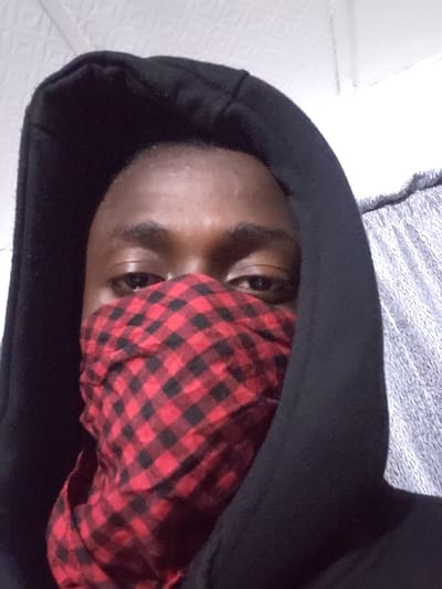 A person wearing a black hoodie and a red and black checkered bandana covering the lower half of their face. Their eyes are visible above the bandana. The background includes a textured white wall and a gray, possibly metallic, surface. The person's skin tone is dark. The image appears to have been taken from a slightly high angle. The photograph is slightly rotated.