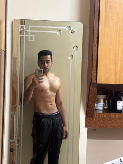 The scene, located indoors in Kolkata, India, depicts a young adult male of South Asian descent taking a mirror selfie. The subject stands shirtless, displaying a muscular physique, and wears black athletic pants with a blue waistband. A golden-colored bracelet is on his left wrist. He holds a silver-colored smartphone in his right hand, directed towards his reflection. The mirror is large, rectangular, and features etched geometric patterns with multiple circular reflective studs. To the left of the mirror, a wooden wardrobe door with a silver handle and a hanging keychain is partially visible. To the right, a brown wooden shelving unit includes an open shelf displaying a white tub labeled "ATINE HCL," various dark bottles, and one black bottle partially identified as "creed." The walls are light-colored.