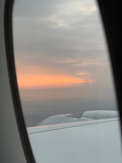 The image, taken from an airplane window, captures a dramatic sky scene, likely at sunrise or sunset, above the Baiyun District, China. The dark frame of the aircraft window outlines the view. To the right, a significant part of the airplane's wing and engine cowling is visible, detailed with rivets and panel lines in shades of grey and white, subtly reflecting the warm light.
The sky is largely overcast or hazy, with muted grey and white tones in the clouds. In the distance, a faint, darker landmass, consistent with mountains or hills, can be discerned beneath the horizon.
The central focus of the image is a prominent, intense vertical streak of orange and red light, suggesting a powerful light source or reflection. Above this main column, a cluster of a few distinct, bright orange-red lights is visible in the upper mid-distance. The fiery coloration and the distinct vertical emanation strongly suggest a large-scale fire on the ground, possibly a wildfire, with its light and smoke diffused by the atmospheric haze or clouds. There is no clearly legible text visible within the frame.
