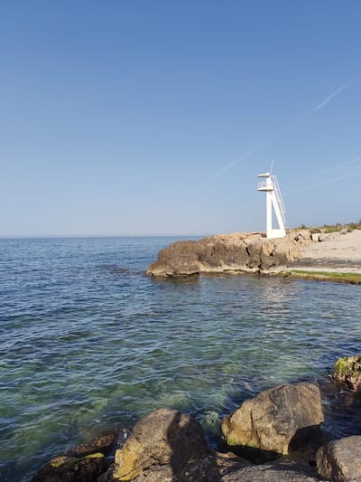 The image depicts a coastal scene in Dénia, Spain. The foreground features a rocky shoreline with clear, turquoise-colored water. Rocks of varying sizes, some covered with algae, are visible in the water and along the edge.
In the midground, a small rocky outcrop extends into the sea. A white lifeguard tower stands on top of the rocks. A narrow, concrete path runs adjacent to the rocks, connecting the area to the beach.
The background consists of a calm, blue sea meeting a clear, blue sky with some contrails. The water appears placid with gentle undulations.