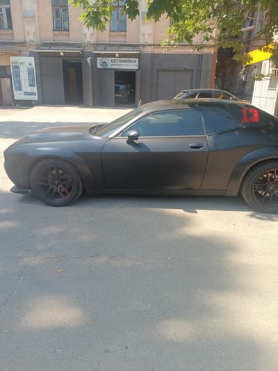 A black matte Dodge Challenger is parked on an asphalt street in front of a building that houses a car wash and detailing service. The car has a red number "13" on its rear quarter panel and red accents on its black rims. The building has a brick facade and several garage doors. One garage door has signage for "Автомийка LeksWash DETAILING" which translates to "Car Wash LeksWash DETAILING." Another sign in Ukrainian, "ПРАЙС-ЛИСТ," which means "PRICE LIST," is displayed on a white board to the left of the building entrance. Green trees with leaves are visible in the upper portion of the image, casting shadows on the car and street. The scene appears to be in a city environment with sunlight illuminating the area.