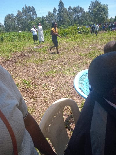 The image was taken at a location in Malava, Kenya, with coordinates 0.529 latitude and 34.813 longitude. The scene is outdoors in a grassy field. There are multiple people present. One person is wearing a white dress and is walking towards the left side of the image. Another person is wearing a yellow shirt and a black skirt, walking towards the right side of the image. There are many other people visible in the background, many of whom are seated in chairs. There is a white plastic chair in the foreground.  The chair is empty. There are many trees in the background, with a blue sky overhead. The scene appears to be a gathering of people for an event.  The ground is covered with grass and dry leaves.