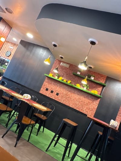 The image displays the unoccupied interior of a commercial establishment, likely a cafe or restaurant, located in Bragança, Portugal. The perspective is an angled, slightly elevated view, looking towards the back and right.
The background features a prominent wall covered in a red-brown faux brick pattern, with a dark, possibly black, wainscoting along its base. Two horizontal, narrow shelves, colored green, are mounted on this brick wall. The lower shelf holds multiple yellow, flower-like decorative items and small potted plants, while the upper shelf similarly displays potted plants and some yellow decorations. Below these shelves, four black electrical outlets or data ports are visible on the brick.
The ceiling is white with a dark grey/black curved architectural feature running horizontally across the upper portion of the frame. Multiple pendant lights, some with yellow shades and others with dark grey or black shades, hang from the ceiling. Recessed lighting is also visible. A section of the left wall, towards the rear, features dark wood-like paneling and includes a circular clock with Roman numerals. The wall closer to the foreground on the left is solid dark grey or black with a textured surface.
The flooring throughout the main seating area is a bright, artificial green, resembling turf or carpet, contrasting with a darker floor section visible in the extreme foreground-left.
Seating arrangements consist of two types. On the left, a long, narrow wooden table with a reddish-orange runner and placemats is paired with multiple black metal chairs featuring wooden seats. A white, patterned ceramic object rests on this table. On the right, two tall, narrow wooden tables are positioned, each accompanied by black metal bar stools with wooden tops. One of these tables also holds a white, patterned ceramic object.
No individuals are present in the scene.